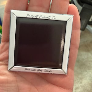 May include: Square magnet with a white border and a black center, held in a hand. The text "Magnet Moments Co." is at the top, and "Moments that Stick!" is at the bottom.