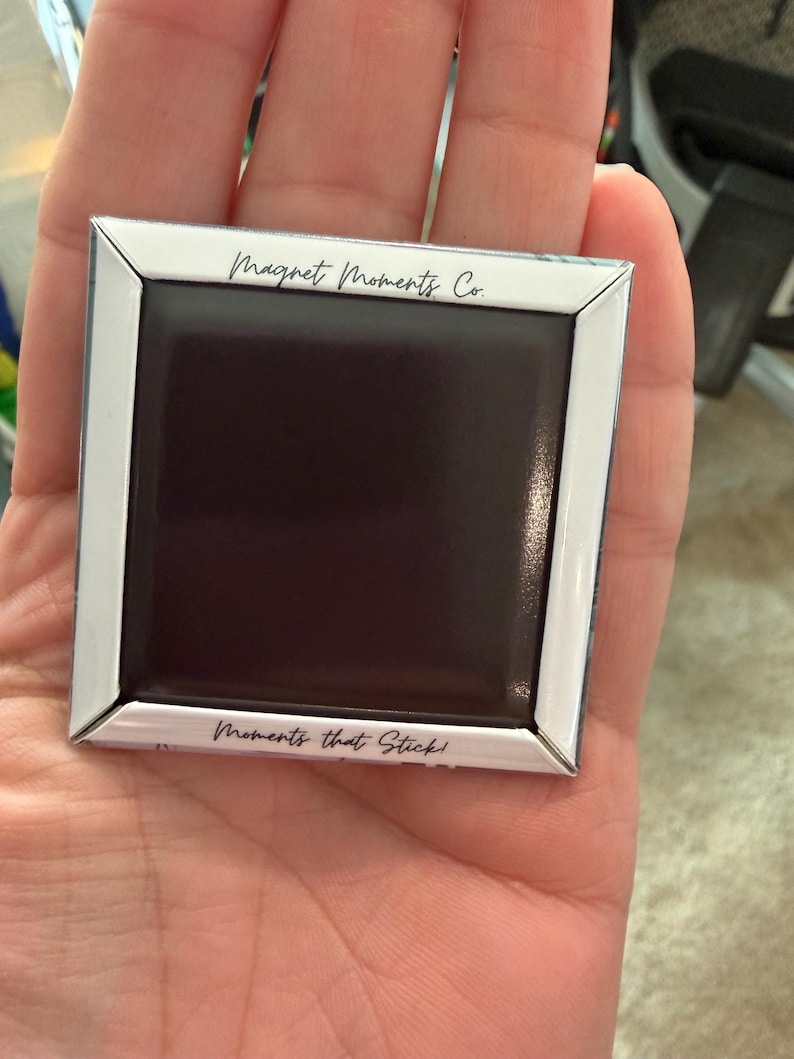 May include: Square magnet with a white border and a black center, held in a hand. The text "Magnet Moments Co." is at the top, and "Moments that Stick!" is at the bottom.