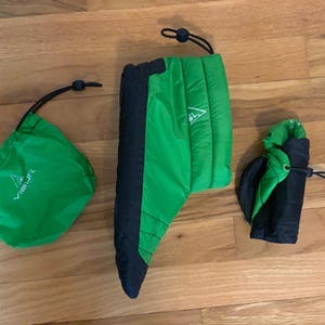 May include: Three pieces of outdoor gear are displayed on a wooden surface. A green drawstring bag, a green and black insulated sleeve, and a folded black and green item with a drawstring closure. Each item has a white triangle logo.