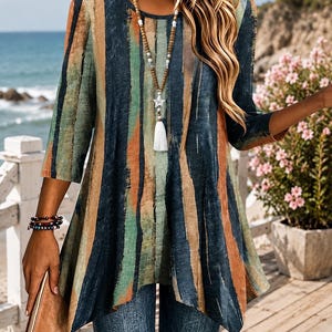 Op de afbeelding: Een tuniek met driekwartmouwen en een verticaal gestreept patroon in marineblauw, saliegroen en oranje. Het kledingstuk heeft een ronde halslijn en een uitlopende zoom. Een lange kralenketting met een sterhanger en een kwastje wordt gedragen.