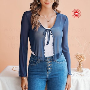 May include: A navy blue sheer long-sleeve bolero featuring a V-neckline with a tie-front closure and ruffled edges. The garment is worn over a white top and paired with blue denim jeans that have a visible button-fly front closure.