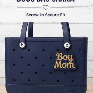 May include: A navy blue Bogg Bag with a gold "Boy Mom" charm. The bag has a rectangular shape with a series of small holes and two handles. The text "Bogg Bag Charm" and "Screw-In Secure Fit" are at the top.