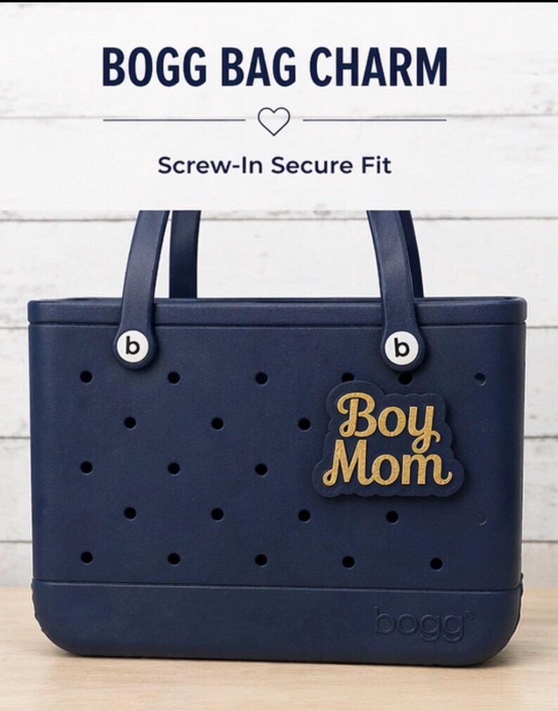 May include: A navy blue Bogg Bag with a gold "Boy Mom" charm. The bag has a rectangular shape with a series of small holes and two handles. The text "Bogg Bag Charm" and "Screw-In Secure Fit" are at the top.