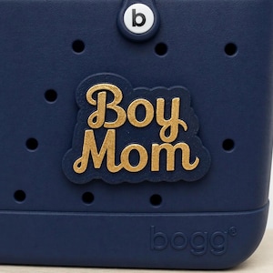 May include: A navy blue tote bag with the words "Boy Mom" in gold lettering. The bag has a circular "b" logo and the brand name "bogg" printed on the bottom. The bag has several small holes.