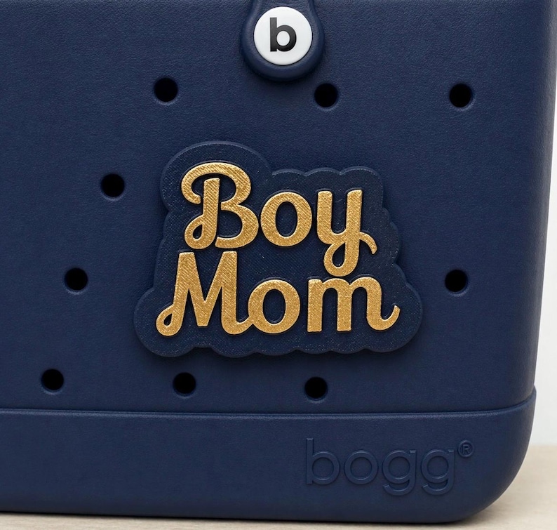 May include: A navy blue tote bag with the words "Boy Mom" in gold lettering. The bag has a circular "b" logo and the brand name "bogg" printed on the bottom. The bag has several small holes.