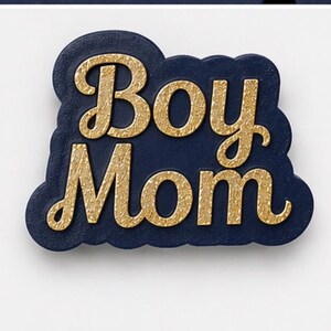 May include: A navy blue and gold-colored pin with the words "Boy Mom" in a decorative font. The pin has a cloud-like shape and is set against a white background.