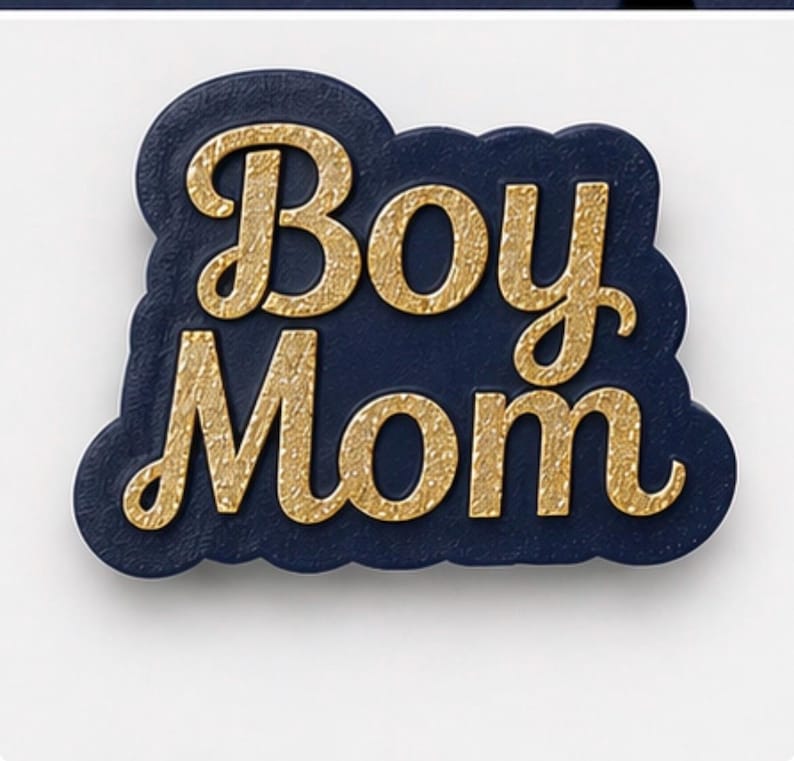 May include: A navy blue and gold-colored pin with the words "Boy Mom" in a decorative font. The pin has a cloud-like shape and is set against a white background.