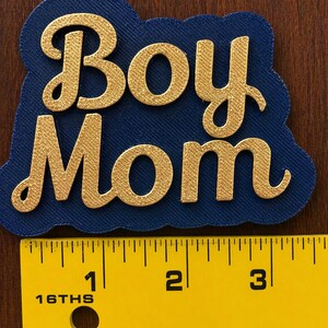 May include: A navy blue die-cut with the words "Boy Mom" in gold script. The text is raised and textured. A yellow ruler with black markings is in the foreground, showing measurements in inches.