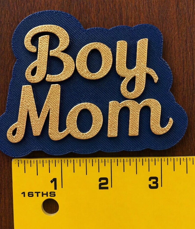 May include: A navy blue die-cut with the words "Boy Mom" in gold script. The text is raised and textured. A yellow ruler with black markings is in the foreground, showing measurements in inches.