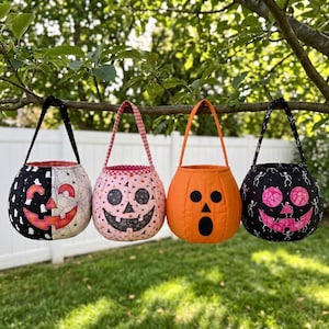 May include: Four fabric pumpkin-shaped treat bags hanging from a tree branch. The first is black and white with a jack-o'-lantern face, the second is pink with a patterned face, the third is solid orange, and the fourth is black with skeletons.