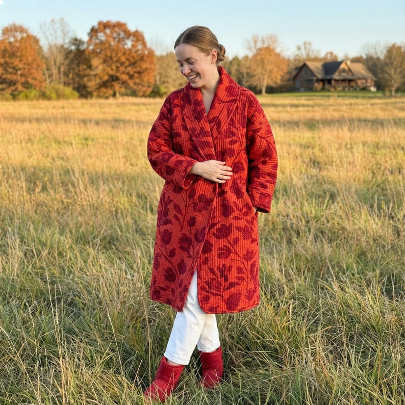 May include: A long, quilted red coat with a floral pattern. The coat is knee-length and has a shawl collar. The person is wearing white pants and red boots. The background is a field with trees and a house.