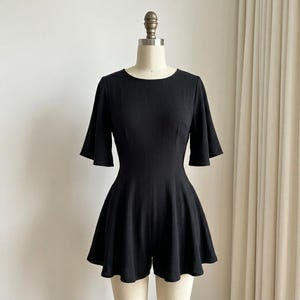 May include: A solid black romper featuring a round neckline, short flared sleeves, and a fitted bodice that transitions into a flared, skirt-like bottom. The fabric has a subtle vertical ribbed texture throughout the entire garment.
