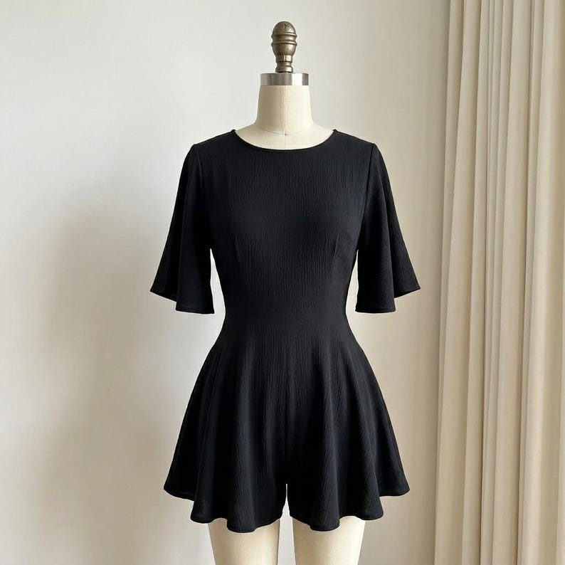May include: A solid black romper featuring a round neckline, short flared sleeves, and a fitted bodice that transitions into a flared, skirt-like bottom. The fabric has a subtle vertical ribbed texture throughout the entire garment.