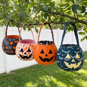 Może przedstawiać: Cztery materiałowe wiaderka na słodycze na Halloween z uchwytami wiszące na gałęzi drzewa. Wiaderka mają różne wzory, w tym pomarańczowe dynie na czarnym tle w pajęczyny, pomarańczowe duszki na białym tle, dynię oraz wzór z nietoperzami.
