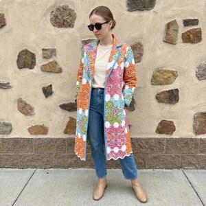 Pu&ograve; includere: Un cappotto lungo e colorato con un motivo paisley nei toni dell'arancione, rosa, verde e bianco. Il cappotto ha un bordo blu e un bordo smerlato. La persona indossa jeans blu, una camicia color crema e stivali color cuoio.