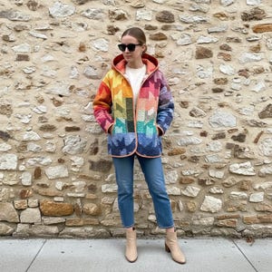Puede incluir: Chaqueta acolchada colorida con un patr&oacute;n geom&eacute;trico de arco&iacute;ris. La chaqueta con capucha se lleva sobre una camiseta blanca y vaqueros azules. Los colores incluyen rojo, naranja, amarillo, verde, azul y morado.