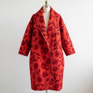 May include: A quilted, knee-length coat in a vibrant orange hue, adorned with a dark red floral pattern. The coat features a shawl collar and long sleeves. The fabric has a ribbed texture, adding depth to the design.