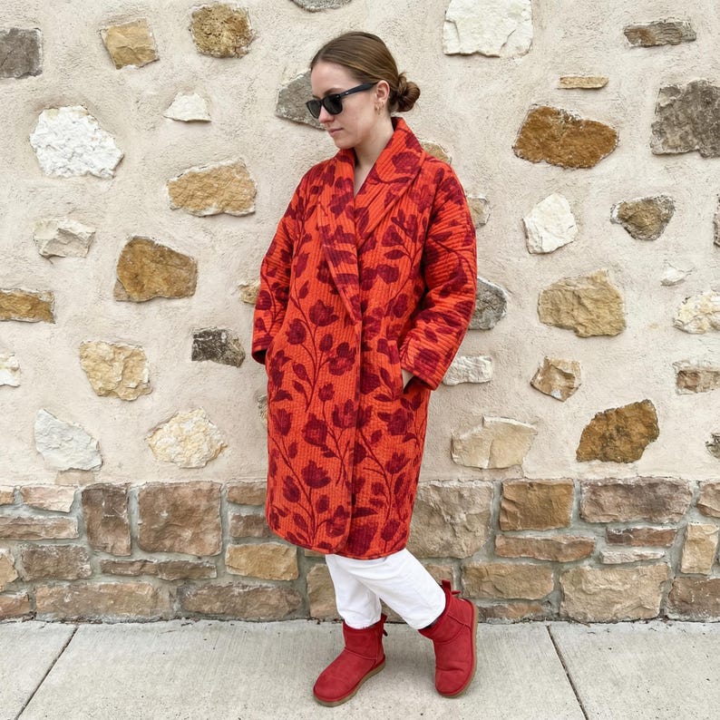May include: A long, red coat with a floral pattern in a darker shade of red. The coat has a shawl collar and pockets. The person is wearing white pants and red boots. The background is a stone wall.