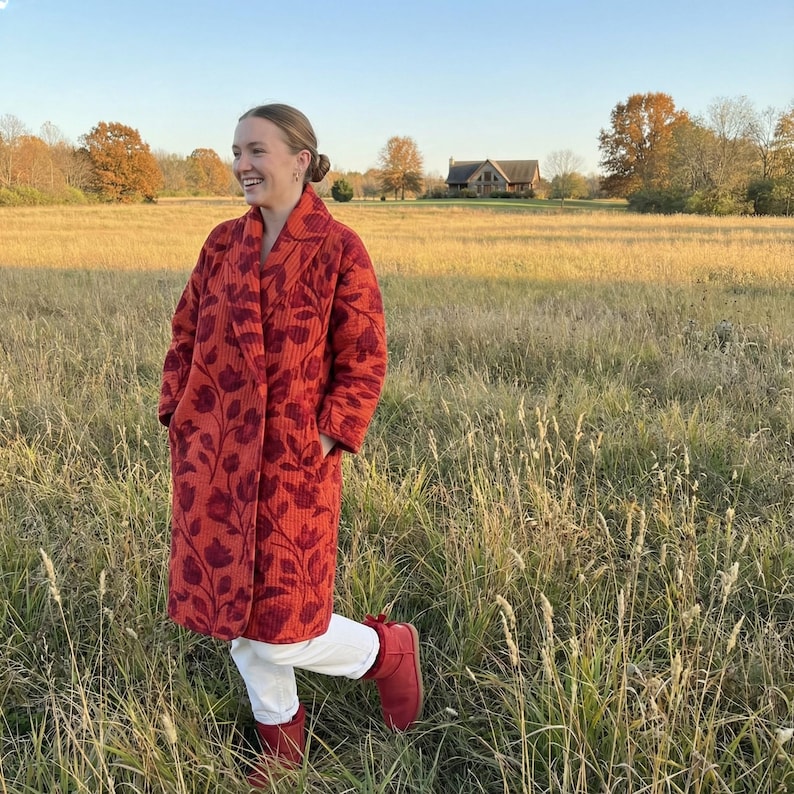 May include: A long, quilted coat in a vibrant red hue, adorned with a floral pattern in a deeper shade of red. The coat features a shawl collar and pockets. The person is wearing white pants and red boots, standing in a field.