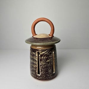 May include: A handcrafted ceramic lantern with a dark brown and beige color scheme. The lantern features a circular handle, a flat top, and a cylindrical body with leaf-like patterns and vertical cutouts. The lantern is approximately 6 inches tall.