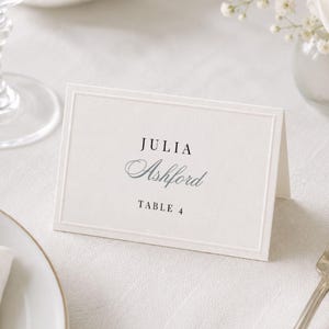 May include: A rectangular, off-white folded place card featuring a thin embossed border. The text printed in black and grey reads JULIA Ashford and TABLE 4. The card is positioned on a white tablecloth next to a silver fork and a knife.