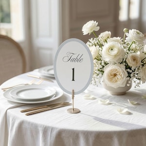 May include: An oval table number card with a light gray border, displaying the word "Table" and the number "1". The card is held by a gold-colored stand. The table is set with white plates, gold cutlery, and a white floral arrangement.