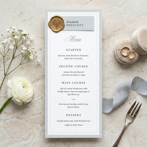 May include: A wedding menu card with a light blue border and the name "Elisabeth Prescott" at the top. The menu lists starter, second course, main course, and dessert options. A gold wax seal and wedding rings are also visible.