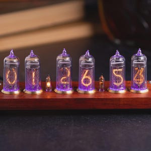 May include: A nixie tube clock featuring six glass tubes with glowing orange numerals and purple base lighting, mounted on a rectangular dark wood base with metallic accents and small screws at the corners, displaying the numbers 0, 1, 2, 6, 5, and 4.
