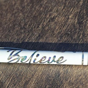 Op de afbeelding: Een witte pen met zilveren details en het woord "Believe" in een holografisch schrift. De pen heeft een zilveren clip en een zilveren punt. Hij ligt op een bruin houten oppervlak.