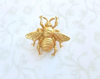 May include: A detailed, golden bee-shaped brooch. The bee has textured wings, body segments, and legs, with its antennae raised. The brooch is set against a white background with a subtle floral pattern.
