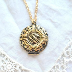 May include: A gold-colored sunflower pendant necklace. The pendant features a detailed sunflower design with a textured center and petals. The necklace has a delicate gold chain and is set against a white background.