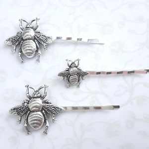 May include: Three silver-colored bee hair pins. Each pin features a detailed bee charm with wings and body segments. The pins are on a white background with a subtle floral pattern, showcasing the hair accessories.