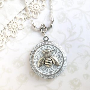 May include: A silver pendant necklace with a bee motif. The circular pendant has a white, textured background and a raised bee design. The necklace features a fine chain and a decorative clasp, suitable for everyday wear.