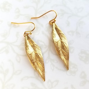 May include: A pair of gold-colored leaf-shaped earrings. The earrings have a textured surface with detailed vein patterns, and are suspended from gold-colored ear wires. The earrings are displayed on a white background with a subtle floral pattern.