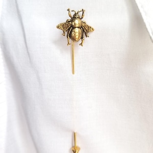 May include: A gold-colored bee-shaped lapel pin. The pin features detailed wings and body, with a long, slender pin extending from the bee's body. The pin is shown on a white fabric background.