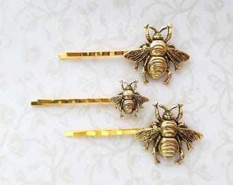 May include: Three gold-colored bobby pins with bee-shaped embellishments. The bees have detailed wings and bodies. The bobby pins are on a white background with a subtle floral pattern.