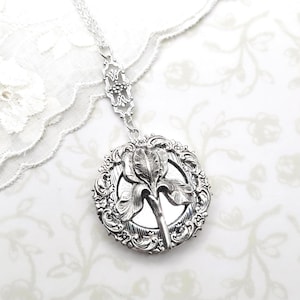 May include: Silver-toned pendant necklace featuring a detailed iris flower design within a round, ornate frame. The pendant hangs from a delicate chain with decorative accents. The floral design is a prominent feature of this jewelry.