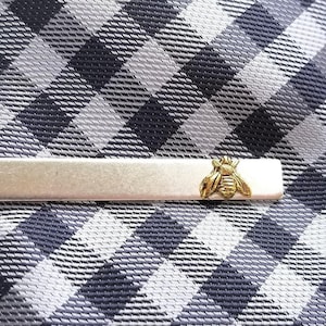 Könnte beinhalten: Eine silberfarbene Krawattenklammer mit einem goldfarbenen Bienenmuster. Die Krawattenklammer ist rechteckig und liegt auf einer schwarz-weiß-grau karierten Krawatte. Die Biene hat detaillierte Flügel und einen Körper.