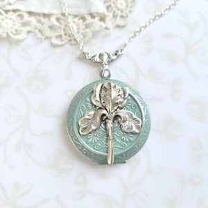 May include: A silver-toned necklace with a round, teal locket featuring a silver iris flower design. The locket has an ornate border and hangs from a delicate chain. The background is a floral pattern.
