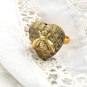 May include: A gold-toned ring featuring a heart-shaped design with a textured diamond pattern. A detailed bee is centered on the heart. The ring is set against a white, textured background.