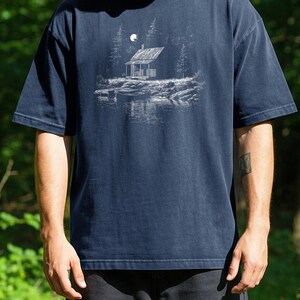 May include: Navy blue t-shirt with a white illustration of a cabin by a lake under a full moon. The cabin is surrounded by trees, and the text "Navy" is printed at the top. The shirt has short sleeves and a relaxed fit.