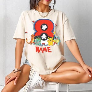 May include: Cream-colored t-shirt featuring a large red and blue number 8 with a black and white circle in the center. Below the number are cartoon characters and the word "NAME" in red.