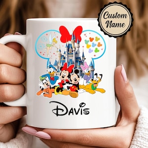May include: White ceramic mug featuring Disney characters Mickey and Minnie Mouse, Donald Duck, Daisy Duck, Goofy, and Pluto. The mug has a castle design, a red bow, and the name "Davis" printed on it. The mug is held by a person.