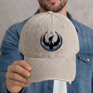 May include: A beige baseball cap featuring a black bird emblem with a blue and white design. The cap has a curved brim and a classic six-panel construction. The bird emblem is centered on the front of the cap.