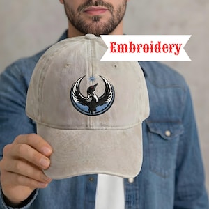 May include: A light beige baseball cap with a circular embroidered design featuring a black bird with outstretched wings, a blue border, and a star. The word "Embroidery" is written in red across the top of the cap.