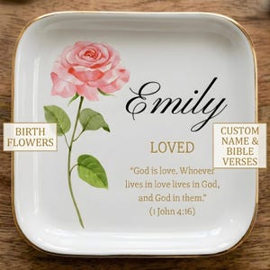 May include: A square white ceramic trinket dish with a gold-trimmed edge. The dish features a watercolor illustration of a pink rose with green leaves. It is personalized with the name Emily, the word LOVED, and the Bible verse 1 John 4:16.