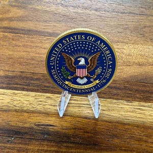 May include: A commemorative coin with a blue and gold rim, featuring the words "United States of America" and "Semiquincentennial 1776-2026." The coin displays an eagle with a shield and is displayed on a clear plastic stand.
