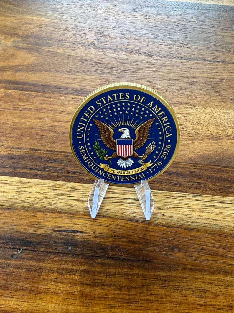 May include: A commemorative coin with a blue and gold rim, featuring the words "United States of America" and "Semiquincentennial 1776-2026." The coin displays an eagle with a shield and is displayed on a clear plastic stand.