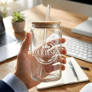 May include: A clear glass can-shaped tumbler with a bamboo lid and a clear glass straw. The tumbler has a white circular design that reads "YOUR DESIGN". The glass is held in a person's hand.