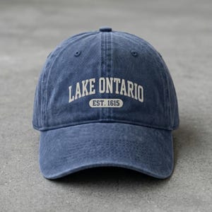 May include: A blue baseball cap with a washed, vintage-style finish. The front features the white text LAKE ONTARIO arched over a smaller rectangular badge containing the text EST. 1615. The cap has a curved brim and a button on the top crown.